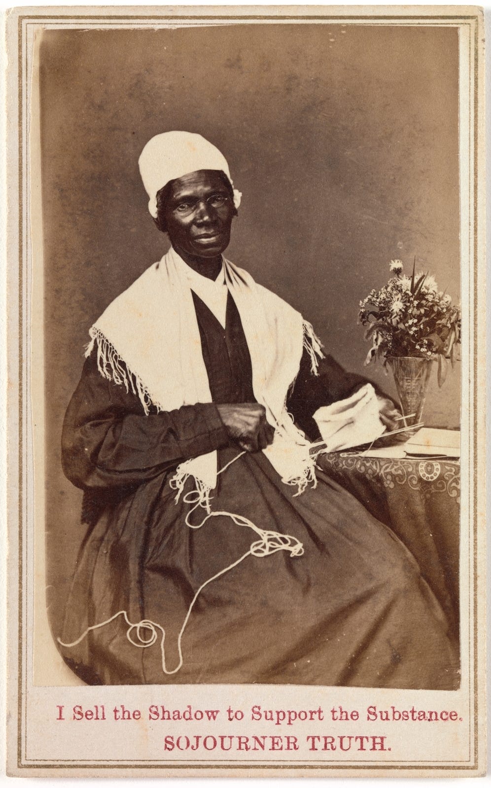 Albumen silver print from glass negative of Sojourner Truth sitting next to a small table with a vase of wild flowers wearing a long black dress, white shawl and cap with a knitting or crochet project in progress in her hands and lap