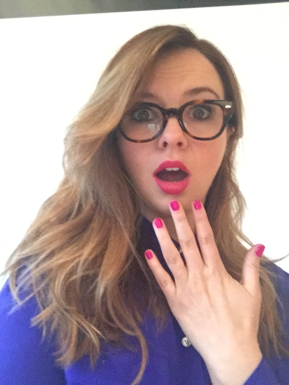 Amber wears reading glasses and holds her hand up to her mouth as if surprised. Her pink nails match her lipstick.