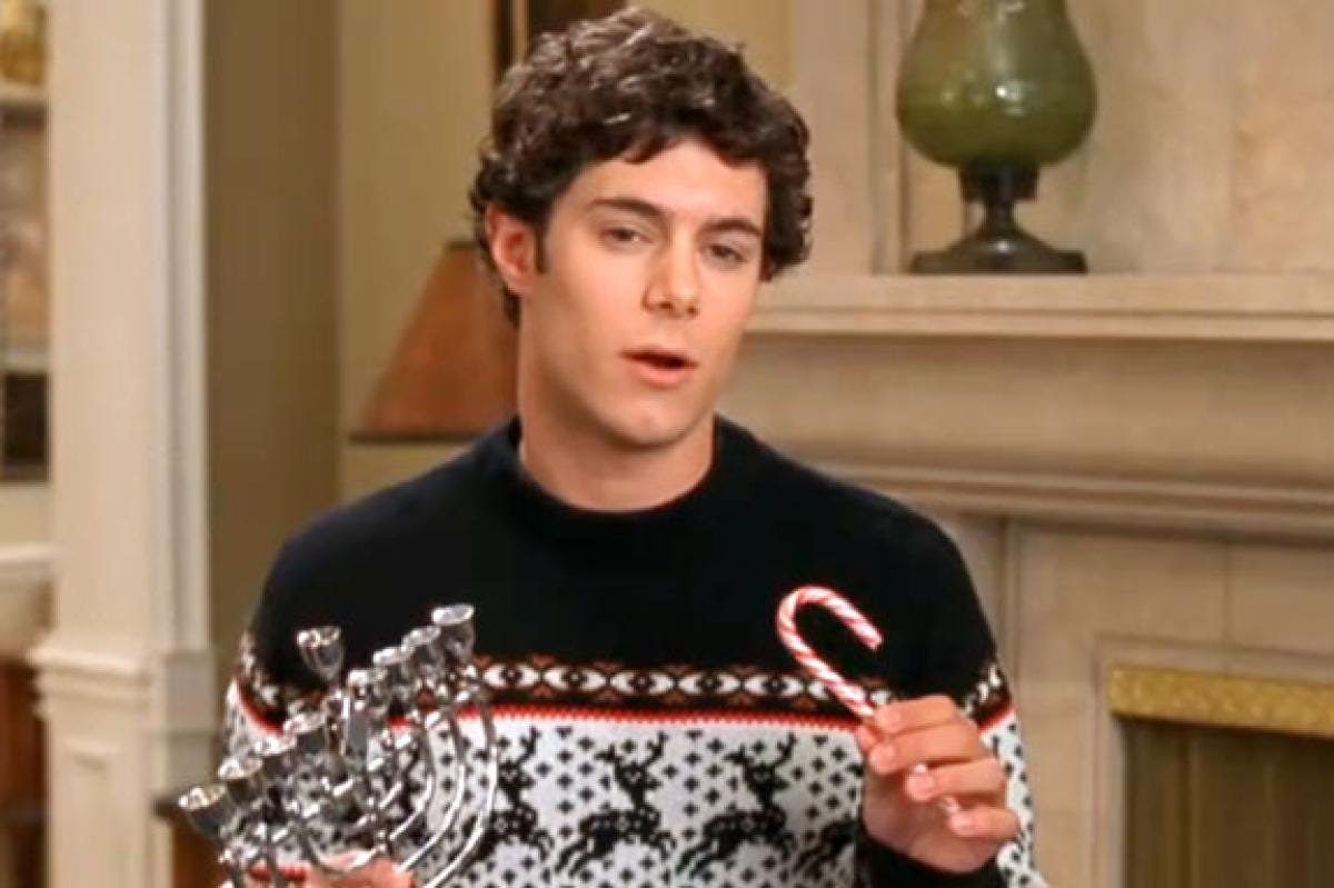 A picture of actor Adam Brody in the television show The O.C. He's wearing an ugly Christmas sweater while holding a candy cane in one hand and a menorah in the other hand.