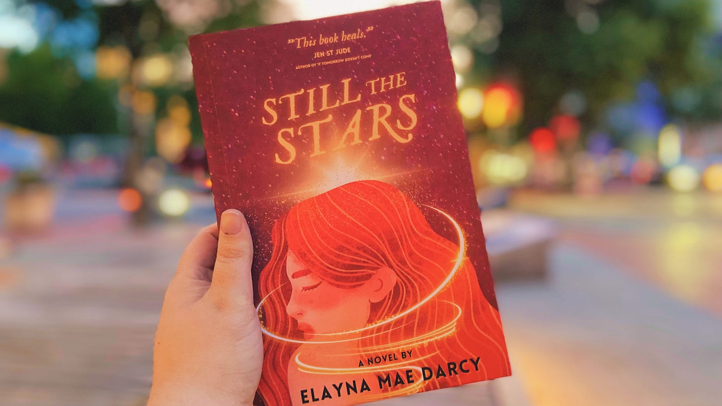 A blurred background of a spring day, with a white hand holding up a copy of STILL THE STARS, which features a vibrant red, orange and gold cover that says the title, has a blurb of "This book heals." from author Jen St Jude, and at the bottom, says, a novel by Elayna Mae Darcy