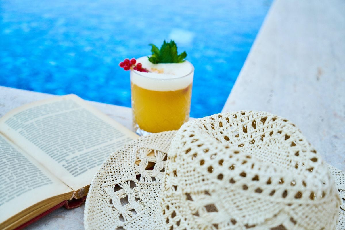 Image of book, summer hat and beverage beside blue swimming pool.