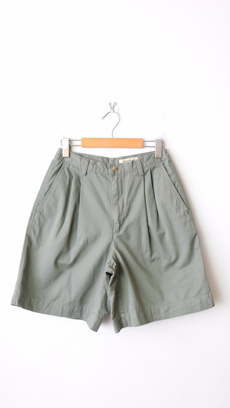 May include: A pair of green cargo shorts with pleats and pockets. The shorts are hanging on a wooden hanger.
