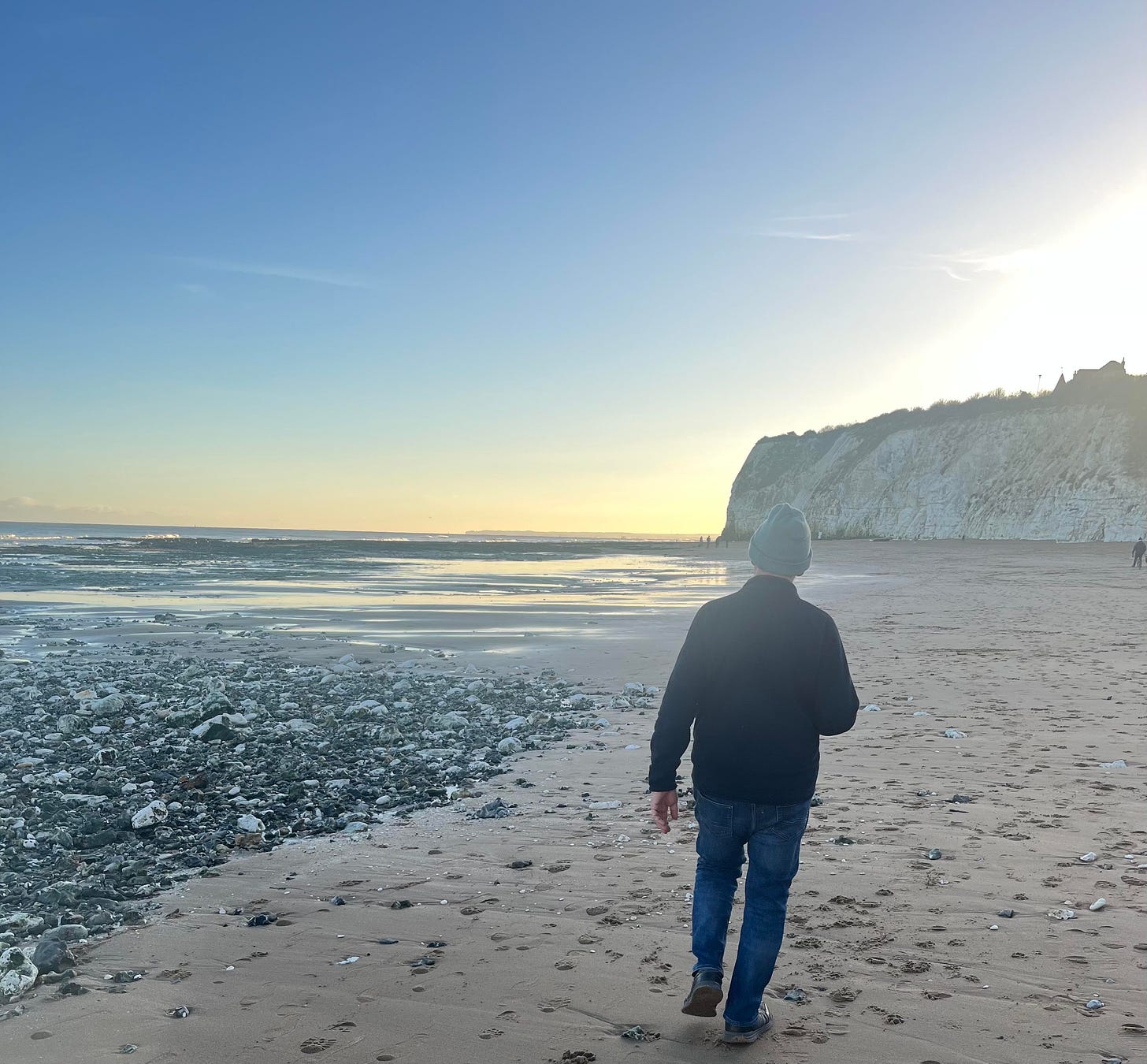 Older white man in knitted cap, jacket, jeans, and rubbers strolls away from the camera on a stone-filled sandy UK coastline.