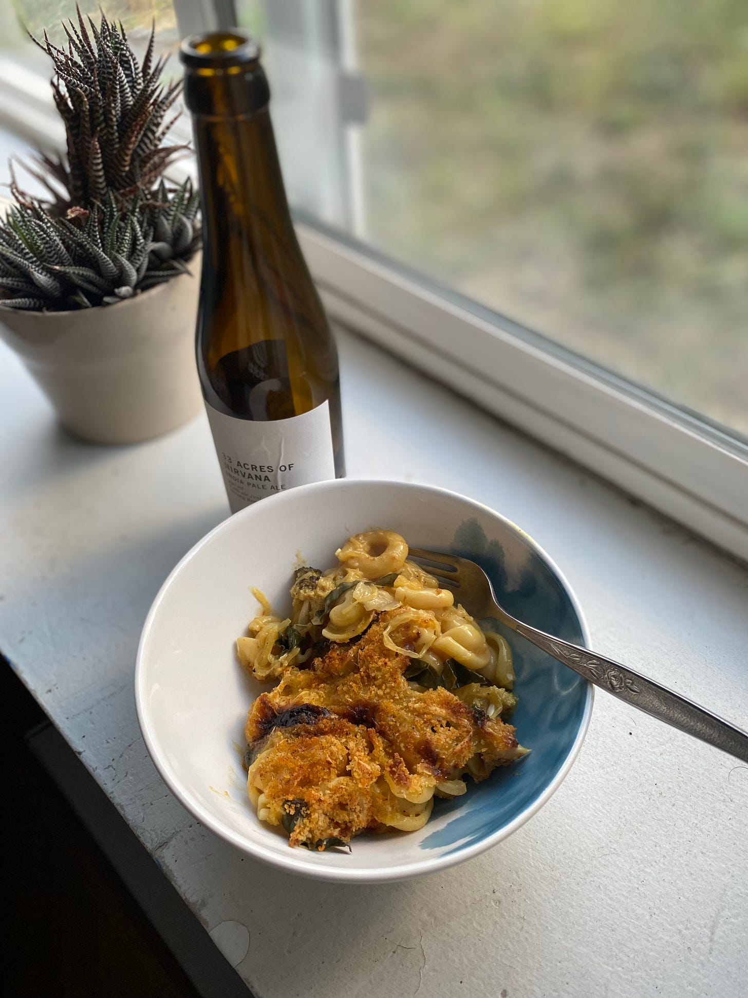 On a windowsill, a white and blue bowl of baked mac & cheeze with kale pieces visible throughout, browned cheese and breadcrumbs on top. Next to it is a bottle of beer, '33 Acres of Nirvana'. Behind them both is a houseplant in a white pot.