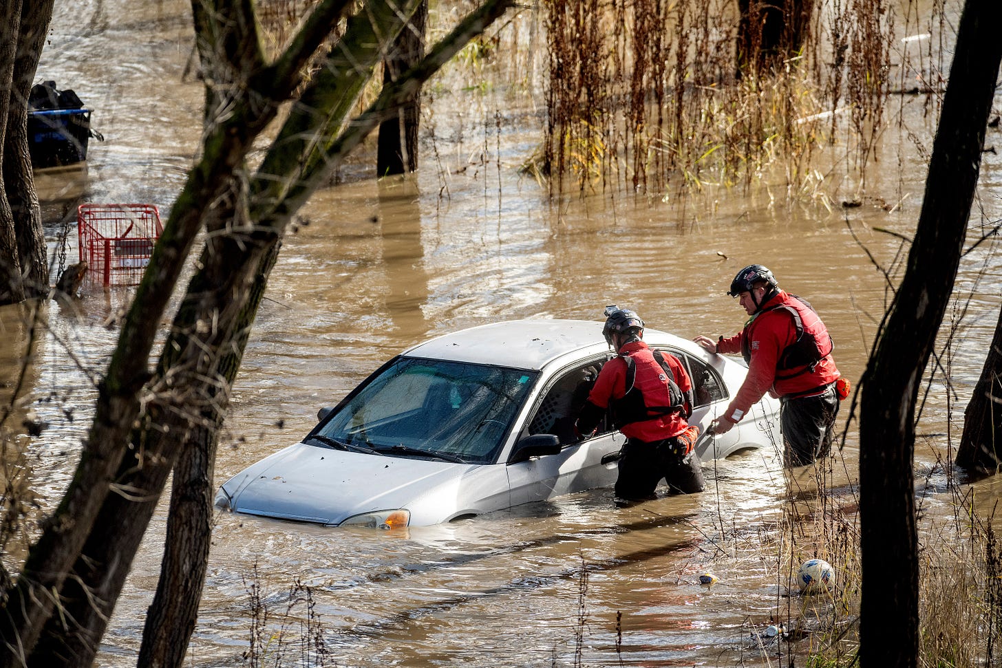 Two search and rescue workers stand by a gray sedan surrounded by floodwater.