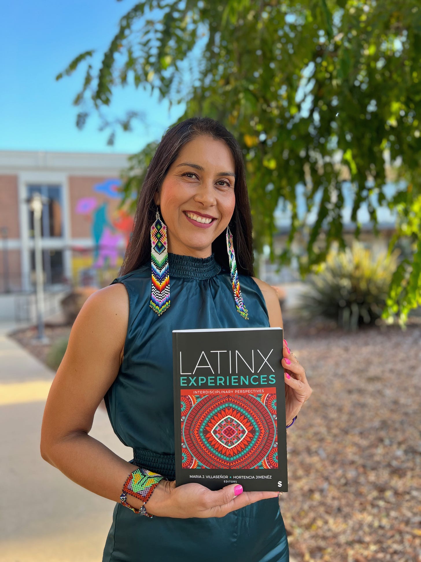 Dra. Hortencia Jimenez stands outside, holding a copy of the book titled Latinx Experiences. She wears colorful, intricate beadwork earrings and a matching bracelet that reflect Indigenous designs, along with a teal sleeveless top. Her long dark hair flows over her shoulders as she smiles warmly at the camera. The background features a building with vibrant murals and lush greenery. 