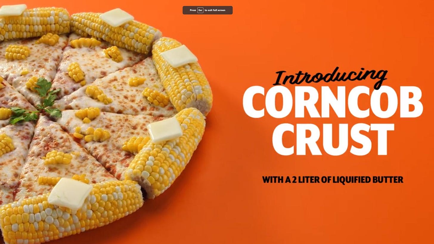A cheese and corn pizza that does appear to have crusts made of whole fresh corns on the cob with squares of butter on them sits against an orange background, with the text “Introducing CORNCOB CRUST, with a 2 liter of liquified butter.” 