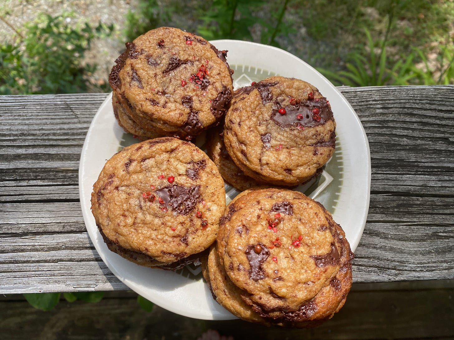 A plate of crinkly chocolate chip cookies sprinkled with pink peppercorns and falky salt, sitting on a porch railing.