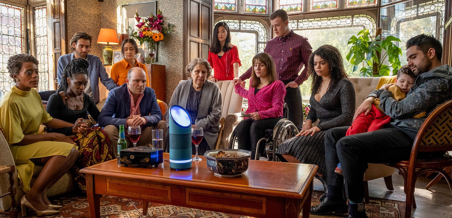 A beautiful old room lined with stained-glass windows is filled with a dozen people, all sitting and standing in a semi-circle, all looking expectantly at a glowing futuristic device sitting on the brown wood coffee table.