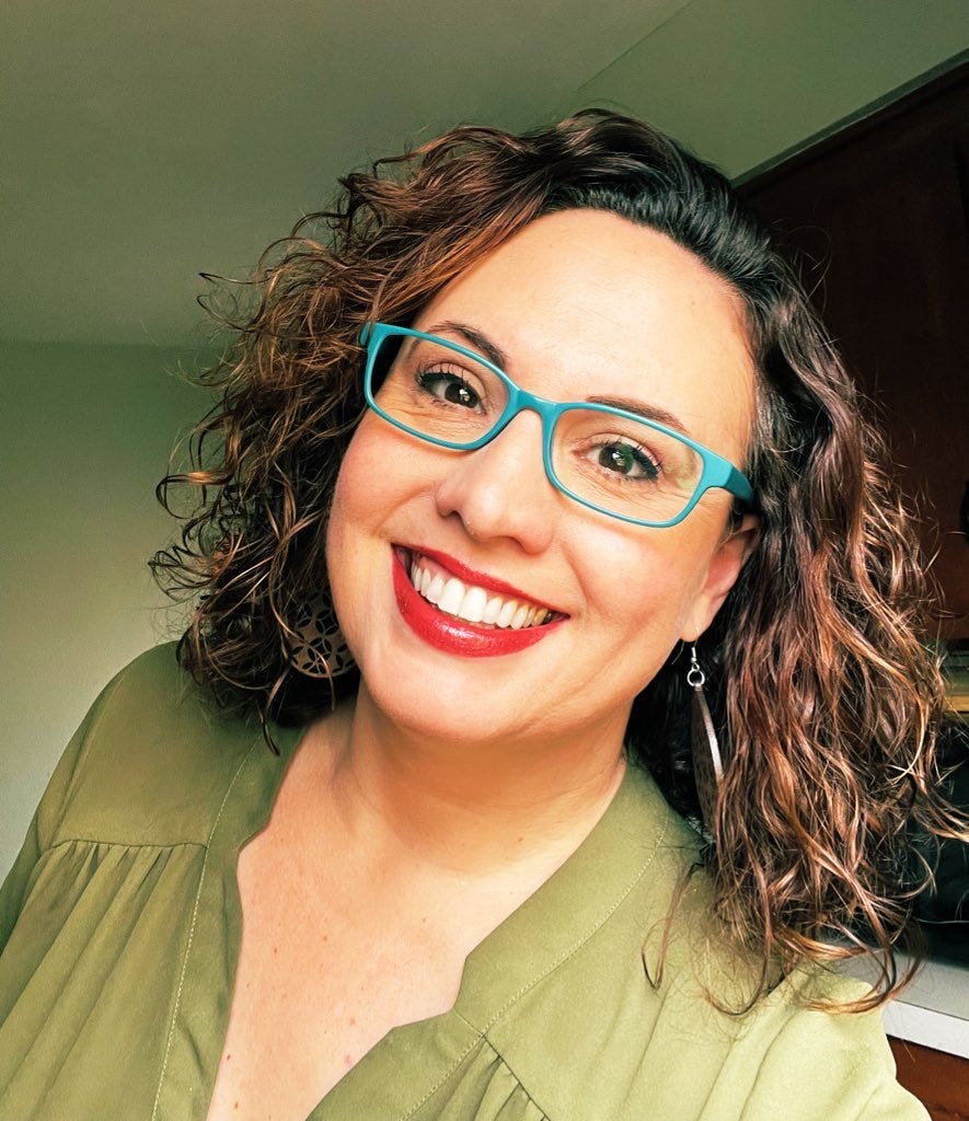 Kristy Acevedo author photo. Woman with brown curly hair and turquoise glasses. Smiling. Wearing a olive green shirt.