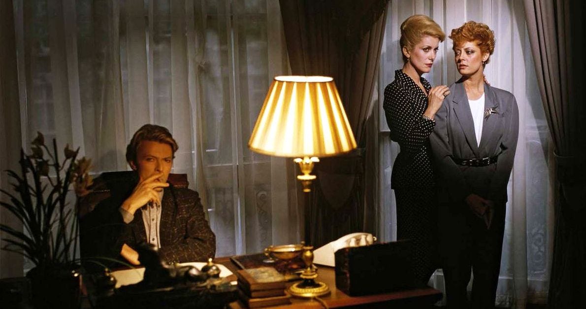 Still from 'The Hunger', a man smokes a cigarette, while two women stand nearby, their body language suggesting intimacy.