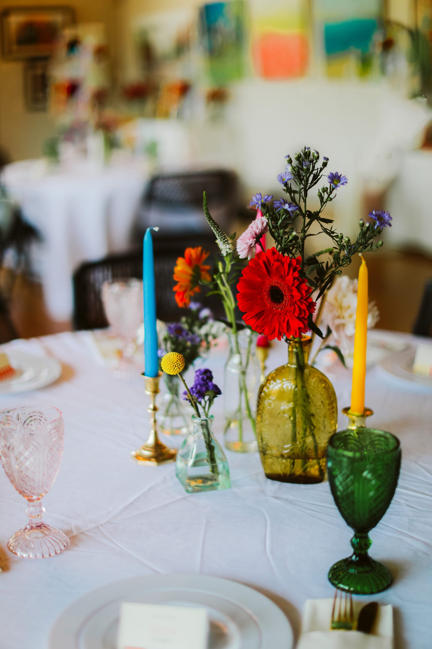 Assortment of bright, wild looking flowers in bright colored vases with multicolored candlesticks on top of a table at a reception.