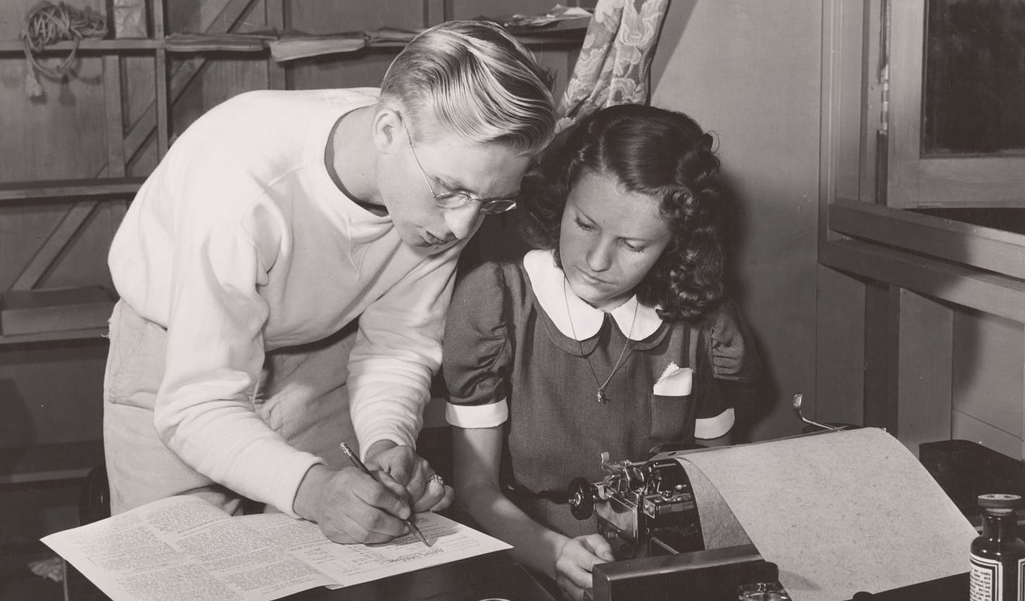 black and white photograph showing an editor standing next to a woman at a typewriter, marking a page while she looks on
