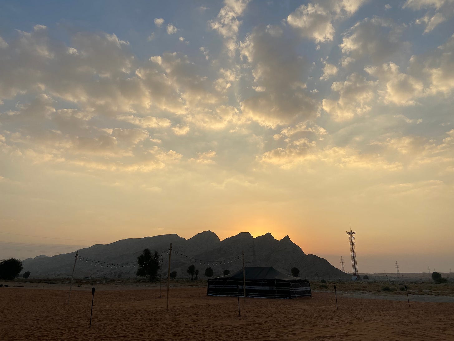 Photo of a sunset in a desert. An Arabic black tent is in the foreground and there a few trees and a mountain in the background.