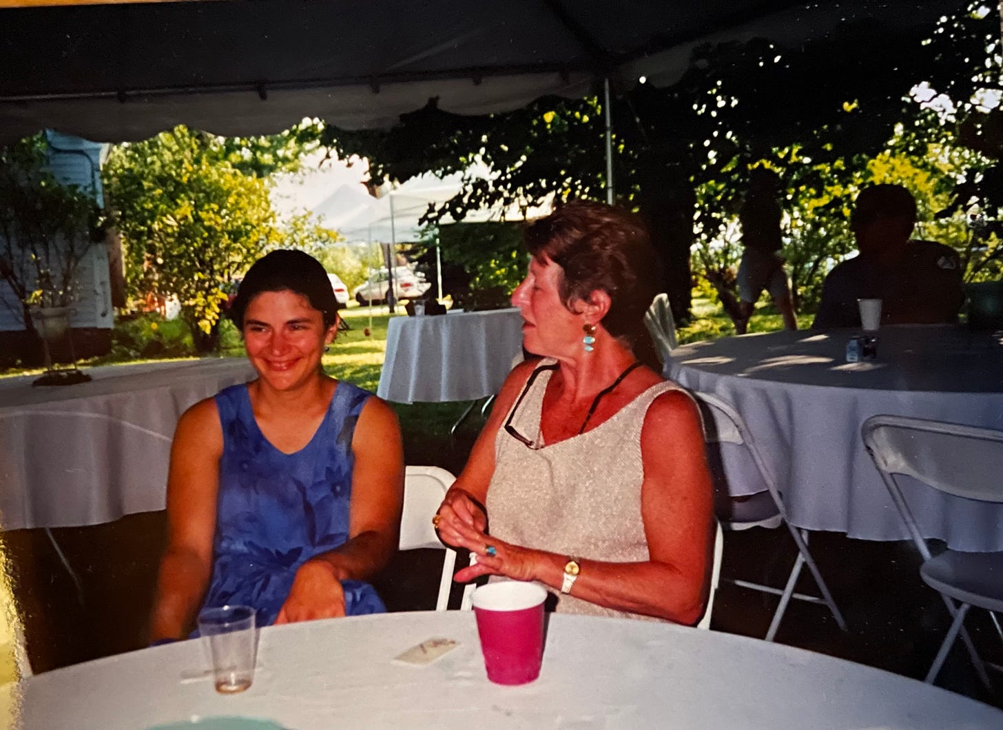 Two women under a even tent speaking at a table with tablecloth, one in younger smiling looking sideways, the other is older turned toward her speaking.