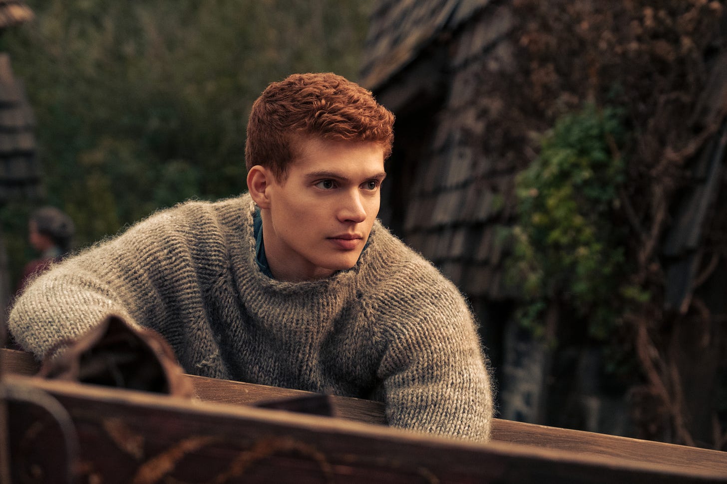 Josha Stradowski as Rand al'Thor in THE WHEEL OF TIME season 1. He has bright red hair, is wearing a light blue handmade sweater, looks comfy as hell, and has no idea of the insanity that awaits him outside of the Two Rivers.