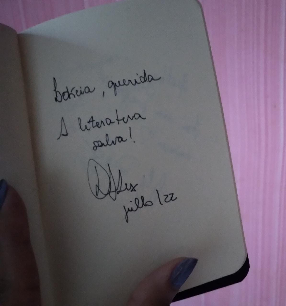 Foto da página de um caderno pequeno, onde se lê o texto escrito em caneta preta: "Letícia, querida: A literatura salva!", seguido por uma assinatura e pela data julho/22.