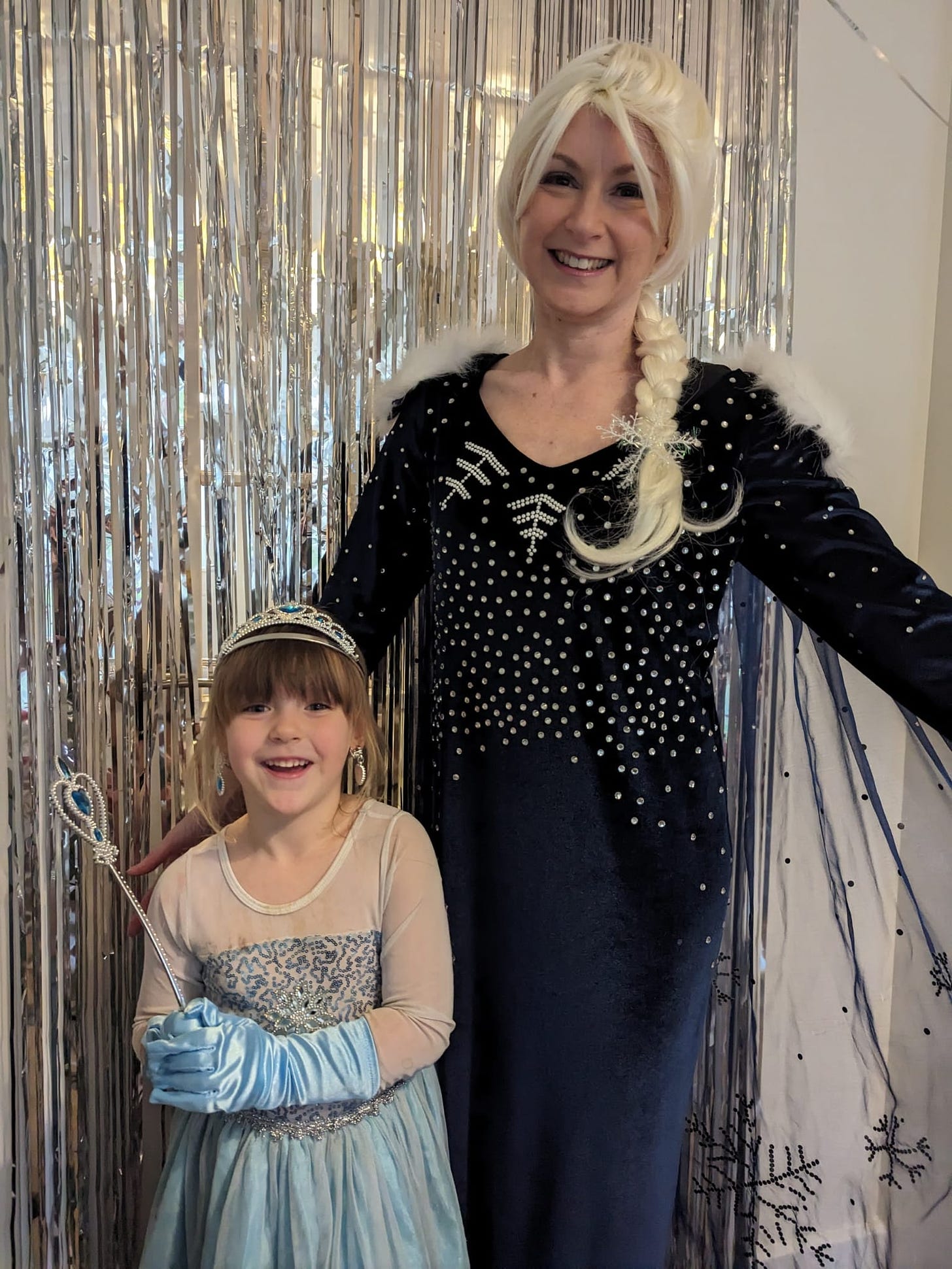 A 5yr old white girl with a brown fringe and blue and white elsa dress and a full real life human elsa from frozen stand in front of a glitter tape curtain