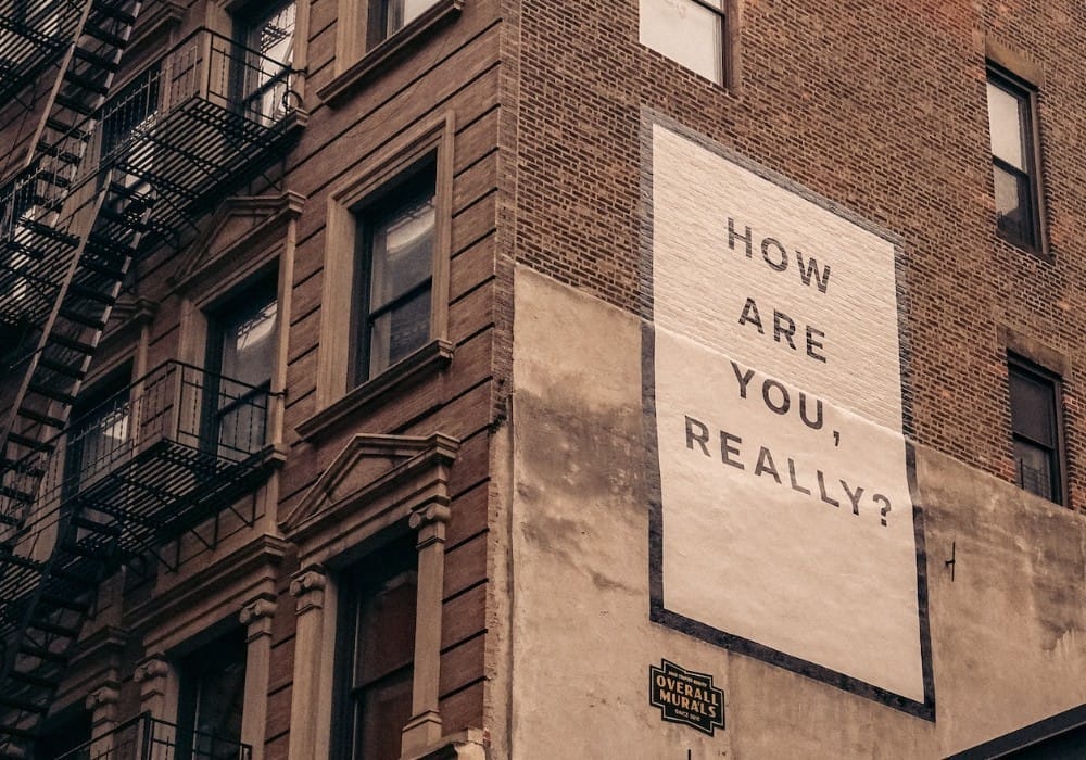 Vintage NY brick building with graffiti on the side saying, "How are you, really?"