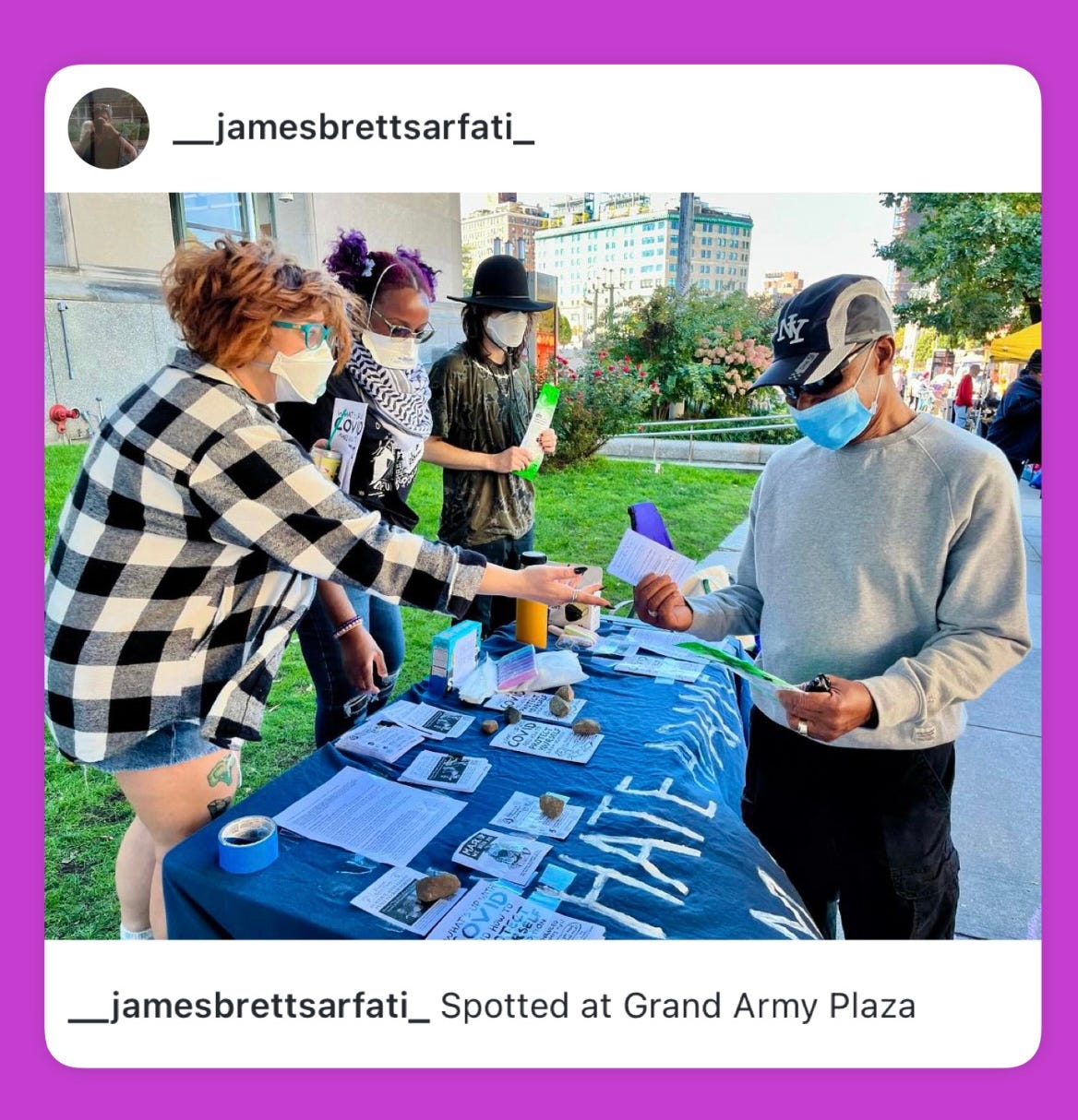 Four people wearing various masks, gathered around a table covered in blue with white text that says, "Hate Adams? Wear a mask!"