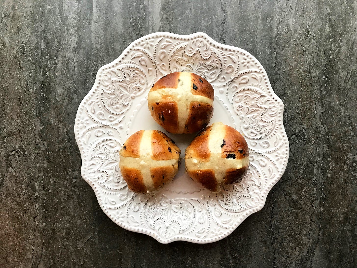 Three baked hot cross buns on an embossed white plate. The buns are a gorgeous umber, speckled with fruit. The paste cross is ivory-coloured in contrast.