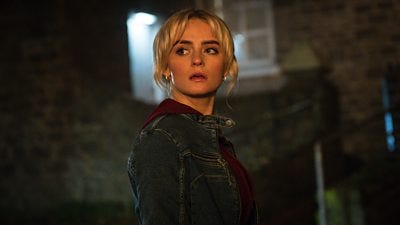 A woman in a denim jacket stands looking at something over her shoulder at night. She looks shocked and distressed.
