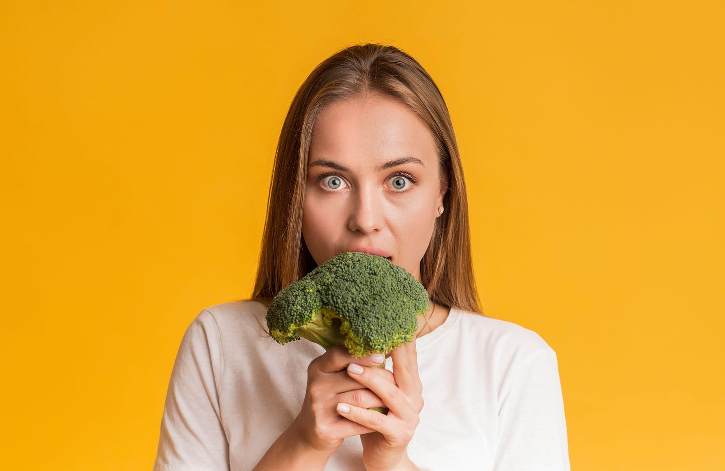 Young woman holding broccoli in front of her mouth infront of a yellow background