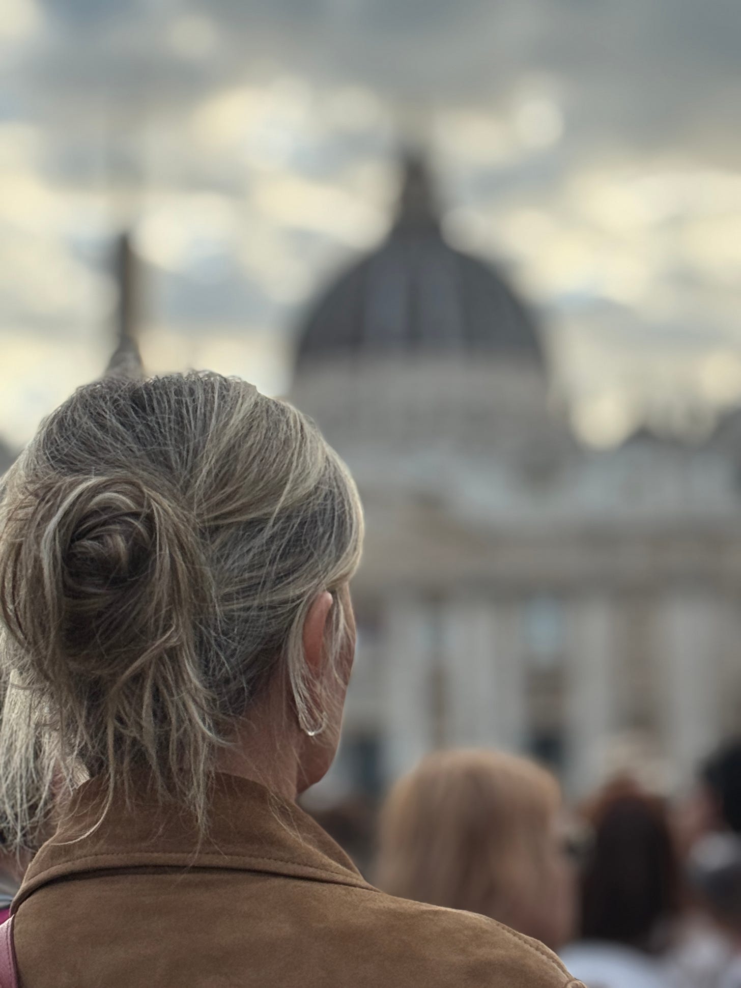 Woman with silver-blonde hair in a loose bun, wearing a brown suede jacket, seen from behind while standing in a crowd at St. Peter’s Square, with the dome of the basilica softly blurred in the background under a moody sky.