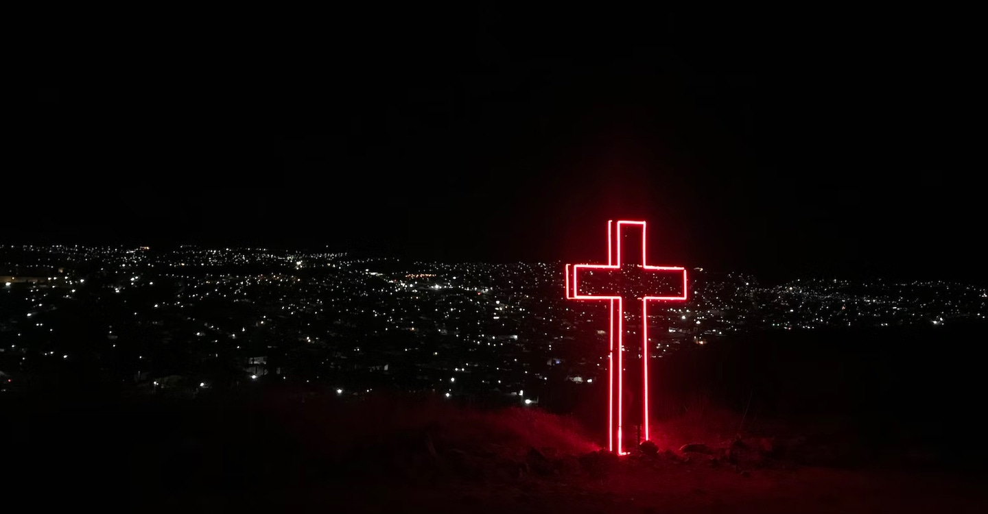A red neon cross stands prominently above a city skyline at night. The image is almost entirely black apart from the cross and the street lights dotted throughout the city.