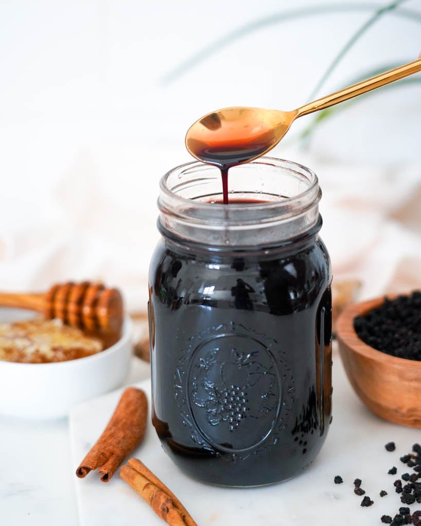 a spoon containing elderberry syrup suspended over a jar of more with honey cinnamon sticks and a small bowl of dried elderberries surrounding it