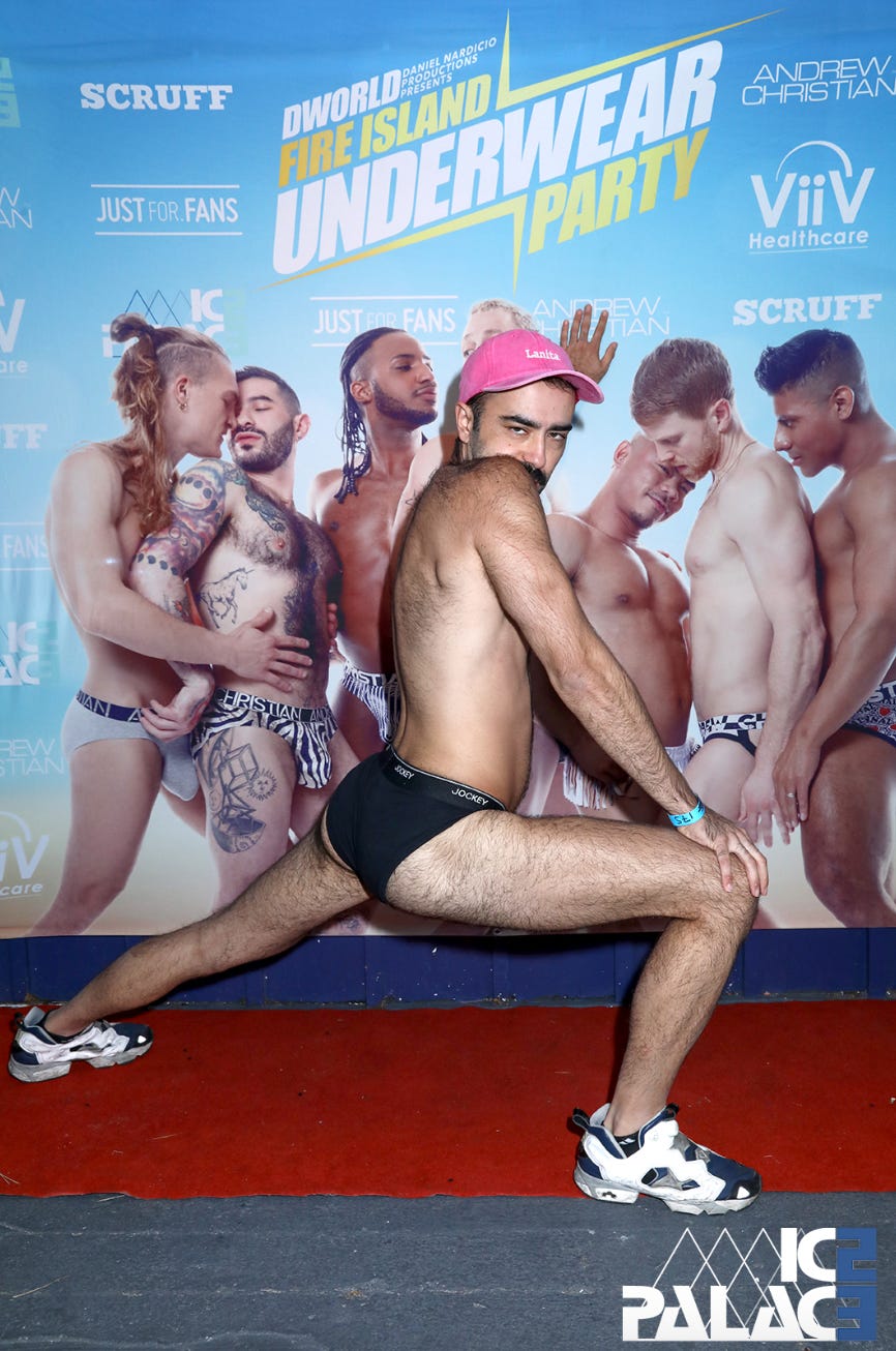 Alt-text: A bearded man in black Jockey underwear, white sneakers, and a pink cap poses dramatically in a deep lunge at the Fire Island Underwear Party. He looks directly at the camera with a playful smirk, one hand on his knee and the other raised in a playful gesture. Behind him, a promotional backdrop featuring muscular men in underwear kissing and embracing is visible, along with logos for Scruff, Andrew Christian, JustFor.Fans, and ViiV Healthcare. The red carpet beneath him adds to the theatricality of the pose. The “Ice Palace” logo is in the bottom right corner.