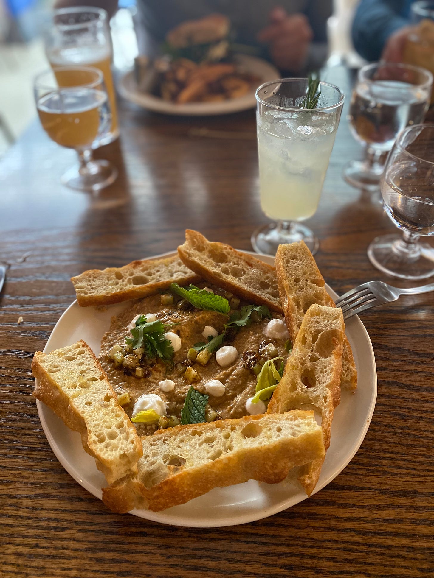 A white plate with the dip described above, surrounded by slices of crusty bread. In the background is a gin and lime soda with rosemary.
