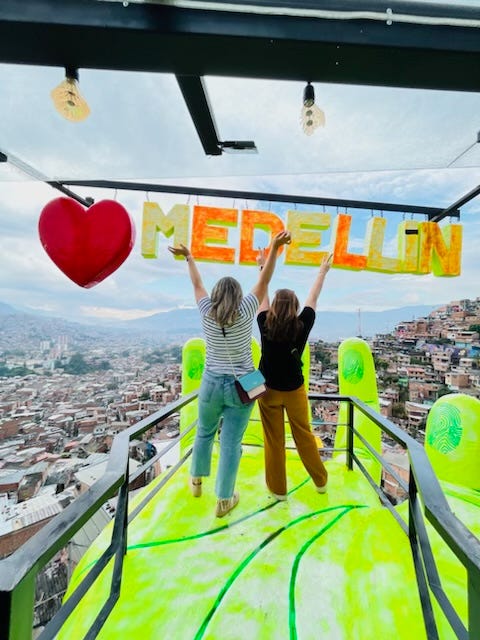 two women standing backs to camera in a giant hand with Medellin sign above