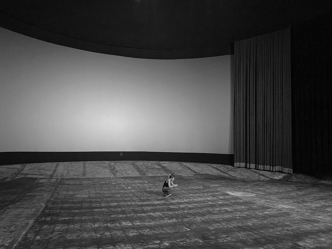 A black and white photo of the interior of the Uptown theater. The curved large screen is in the back with the curtains pulled to one side. The foreground is an empty space where the chairs used to be. A young woman sits squatting on the floor in a contemplative stance.
