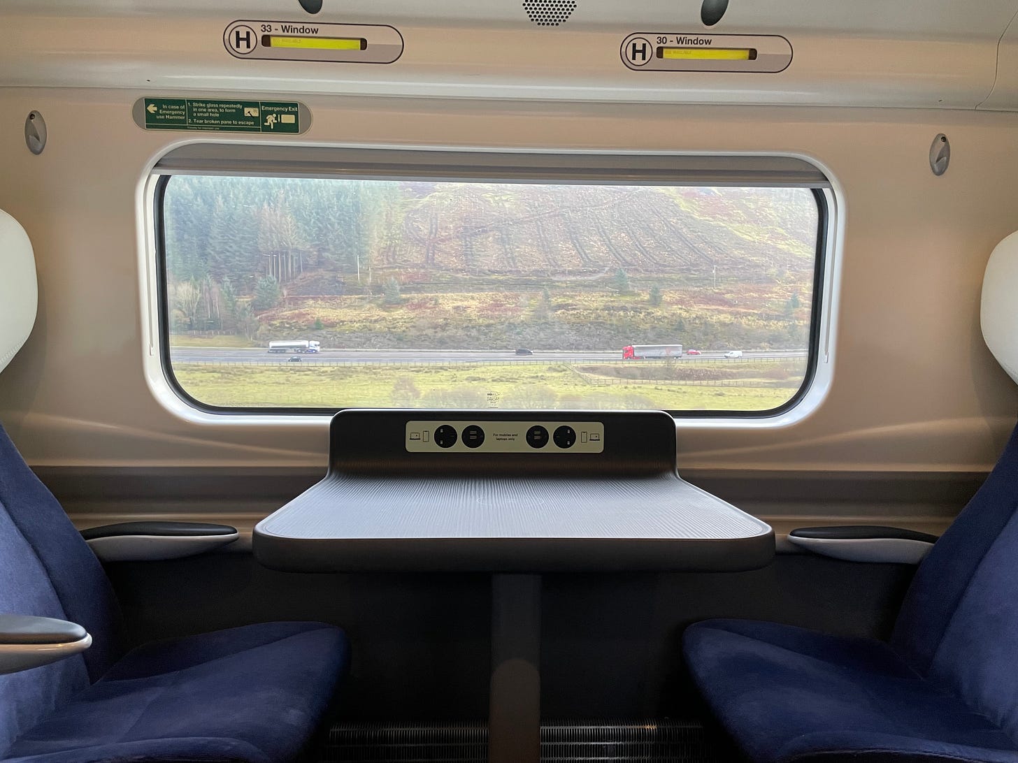 picture looking out of a window of a train, with two seats and a table in the foreground. two lorries and 4 cars are passing on a nearby road