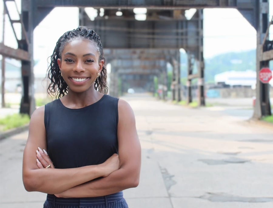 Pennsylvania Representative-elect Lindsay Powell in a campaign photo, standing in an empty street beneath a large iron bridge or elevated railroad, in casual attire, smiling, with her arms crossed