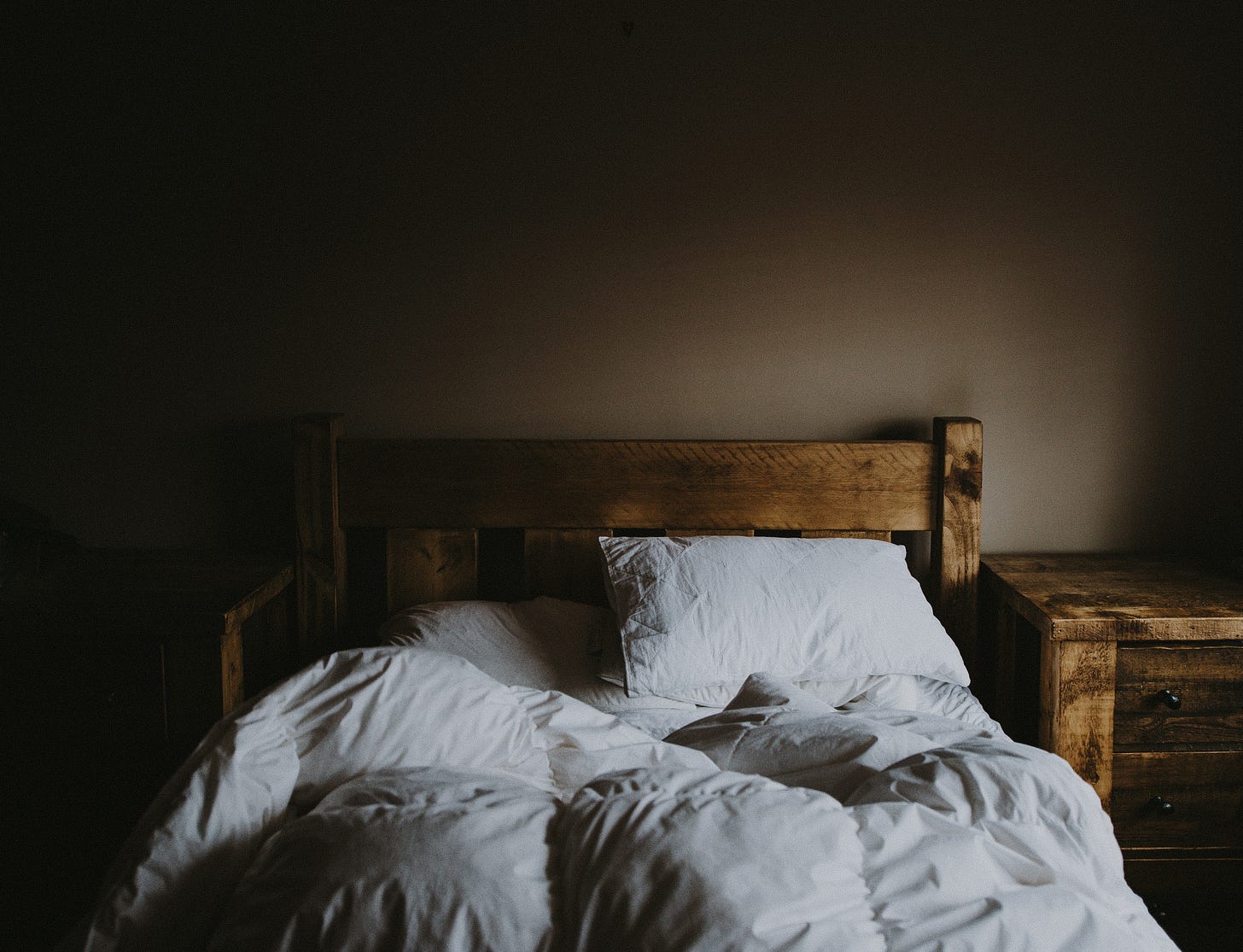 An unmade bed with ruffled sheets in dimly lit by morning light. 