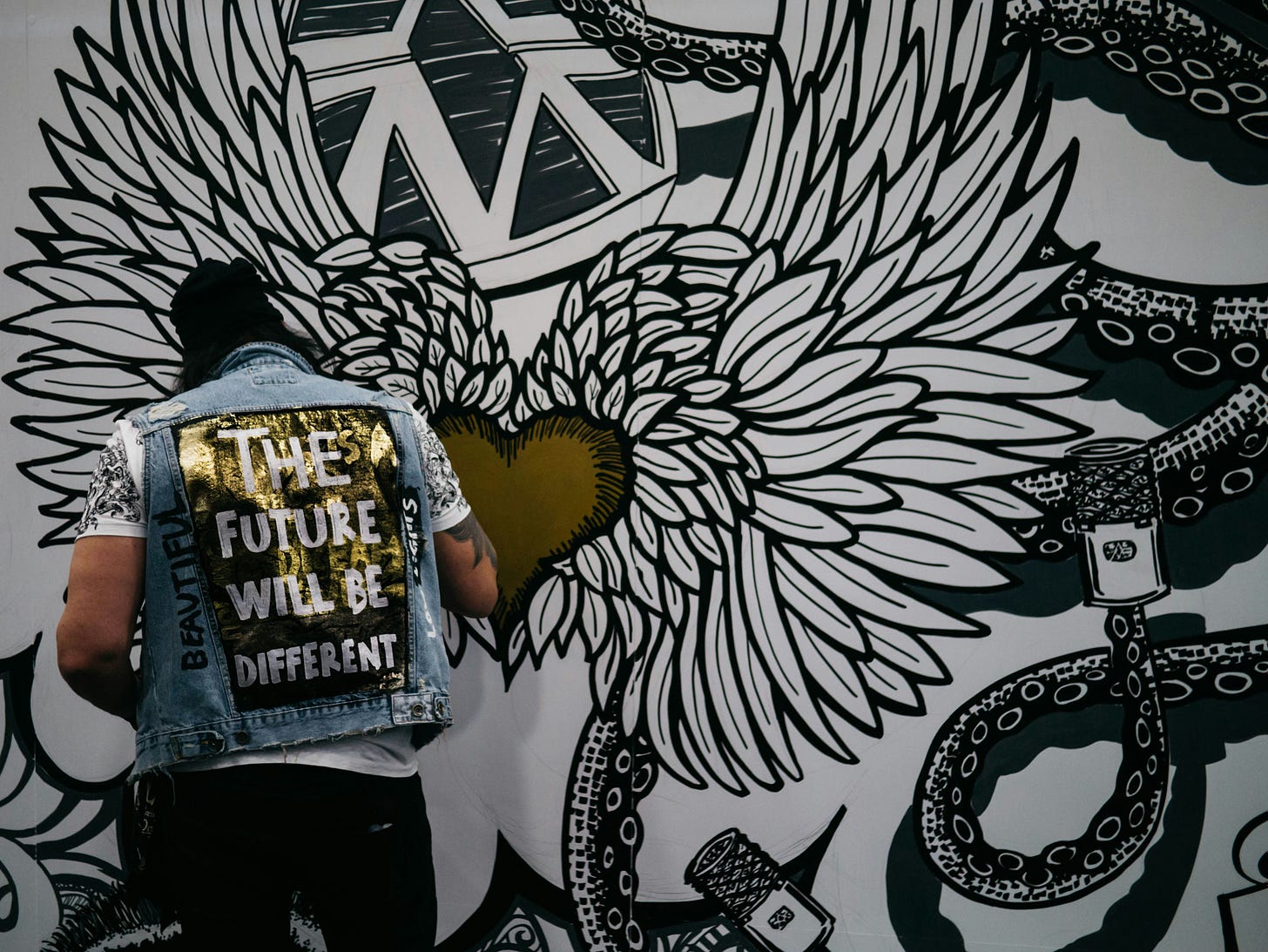 A man with his back to the camera works on a large black, white and gold art installation featuring, at its centre, a winged heart. The man wears a denim vest which reads "the future will be different".