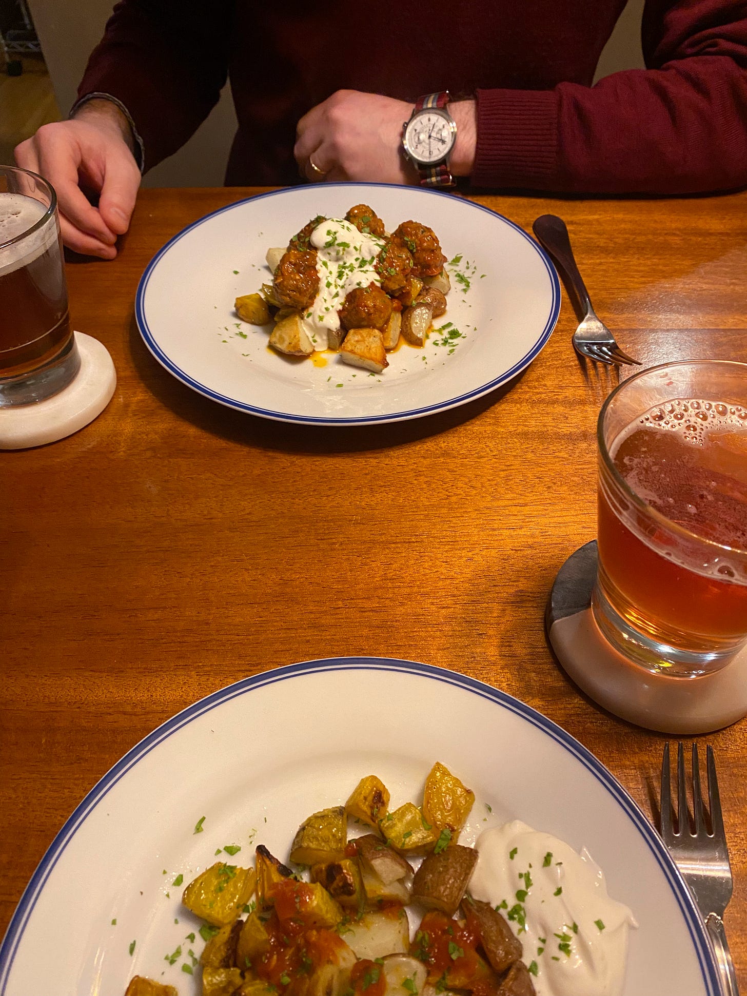 Two plates across from each other on the table, each with roast potatoes, beets, and meatballs in tomato sauce. The dish is garnished with parsley and mint and a dollop of garlic yogurt. Glasses of amber lager sit on coasters at the edges of the plates.