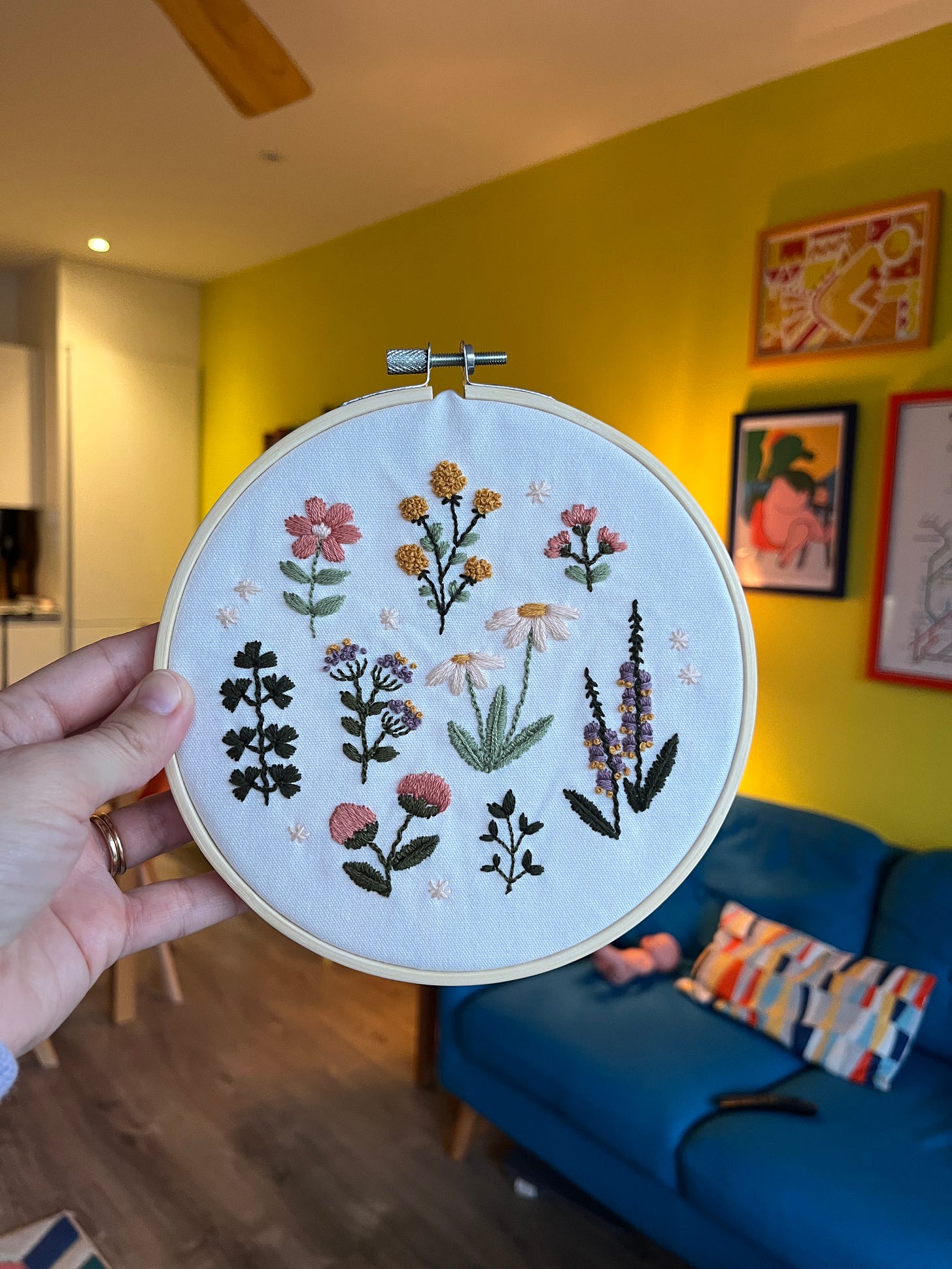 Close up of a few different embroidered flowers all within an embroidery hoop. Hannah is holding this finished piece up in front of her yellow living room wall.