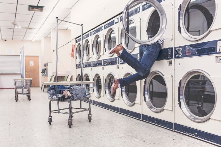A person at a laundromat. Their upper body is entirely in the dryer as they reach for their clothes. Their legs are kicking in the air.