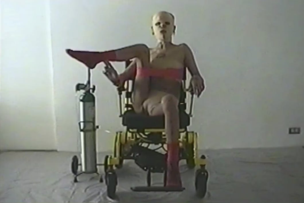 A grainy still, like an older form of film. A disabled person of color rests their leg on an oxygen tank, their leg threaded through an exercise band that doubles as a tube top around their chest. They have bandages covering their face and sit in a wheelchair.