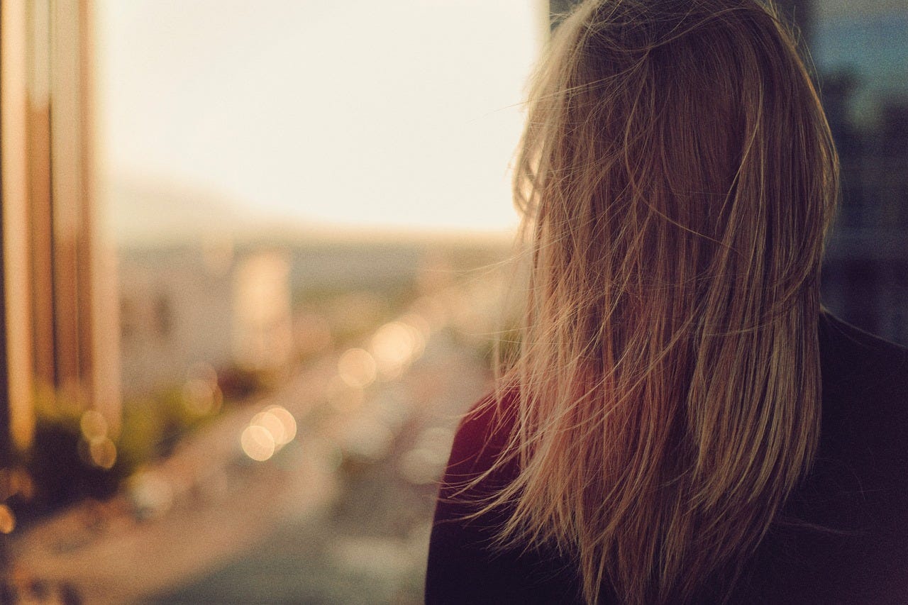 photo of a woman with her back turned to the camera. she seems to be staring out a window or balcony, but the background landscape is out of focus