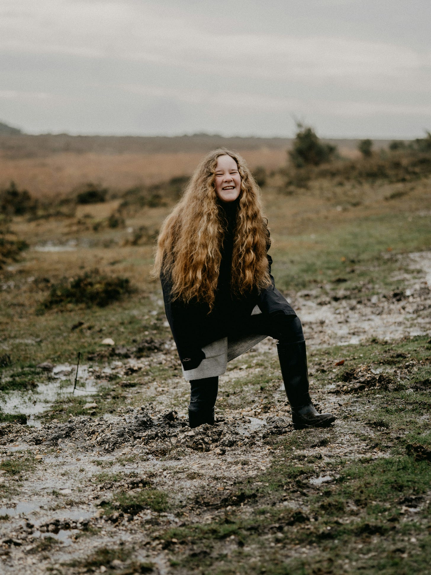 A young person with long, curly hair traversing an unending field during what may be Springtime, as the ground is quite wet. One of their feet is stuck in the mud. They smile, as though mid-laugh and look at the camera.