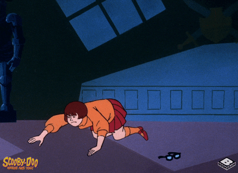 a gif of velma from scooby doo, patting the ground in search of her glasses, which are laying on the ground to her right