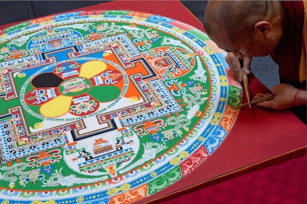 Photo of an intricate sand mandala. To one side a Tibetan monk can be seen carefully adding a minute quantity of colored sand.