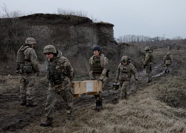 Six soldiers walking in a muddy field, two of them carrying a large wooden box.