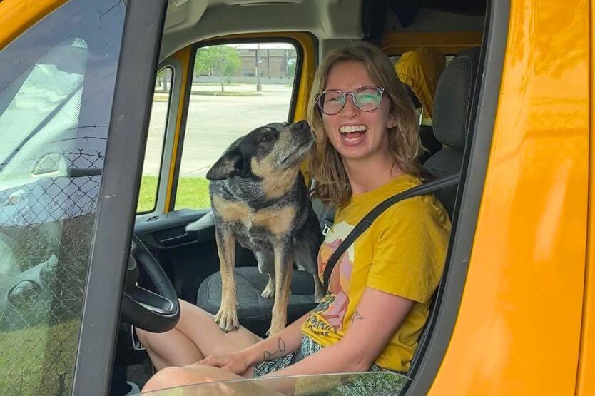 A blue heeler and a young blonde woman sit in the front seat of a yellow van