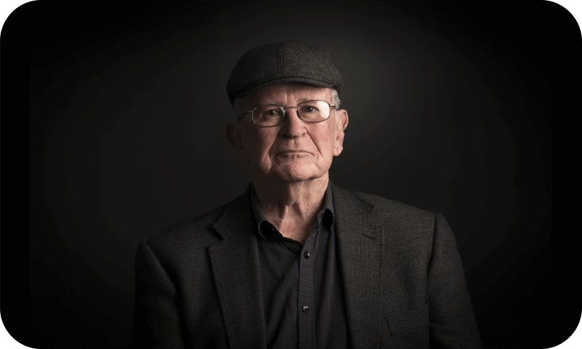 Photographic portrait of an older man in a dark tweed jacket and cheesecutter hat. Beautifully lit so that only his face is really visible. An air of great warmth and cleverness.