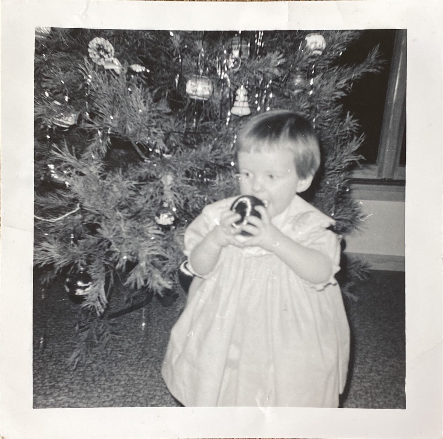 Black and white photo of toddler in front of a Christmas tree, holding satin ornament up to her mouth, as if to bite.